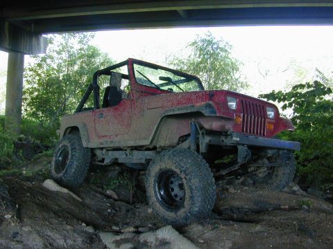 1996 Ford Bronco - A red Jeep Wrangler navigates a rough, muddy terrain beneath an overpass.