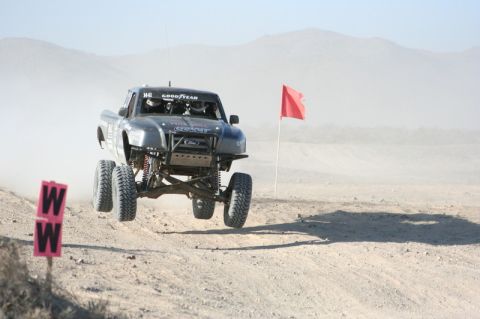 1996 Ford Bronco - A gray off-road truck with Goodyear tires soars through the air during a race, with a red flag marking the course.