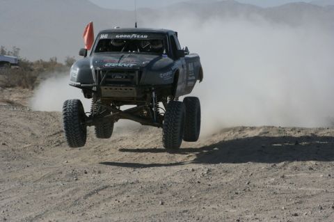 1996 Ford Bronco - Off-road race truck jumps over a dirt mound, kicking up dust.