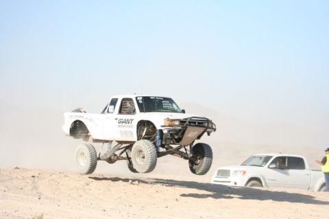 1996 Ford Bronco - A white off-road truck takes flight during a desert race, while another vehicle is stuck in the sand.