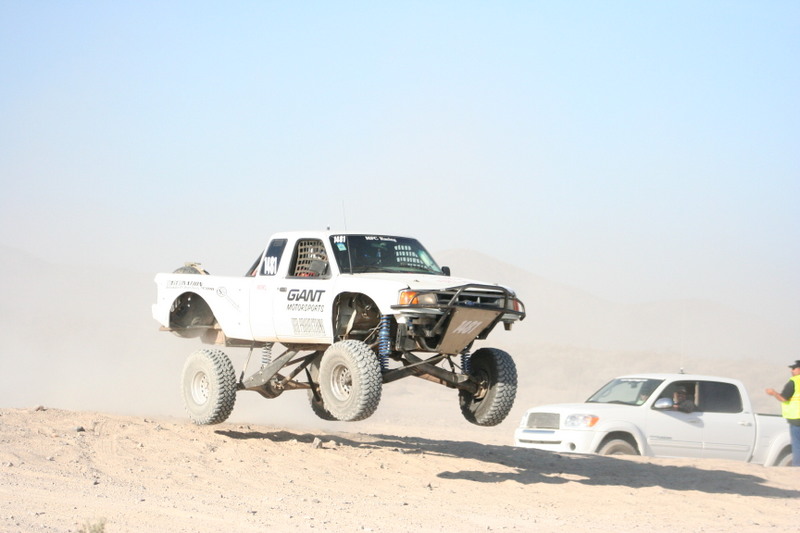 White off-road truck airborne over dirt, another white truck partially buried in sand nearby