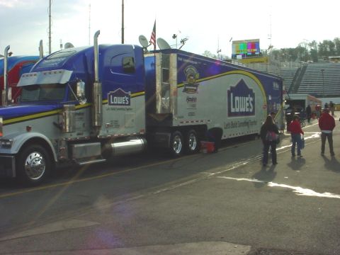 1993 Ford Mustang - A Lowe's branded semi-truck is parked on the asphalt near a race track, with people standing around.