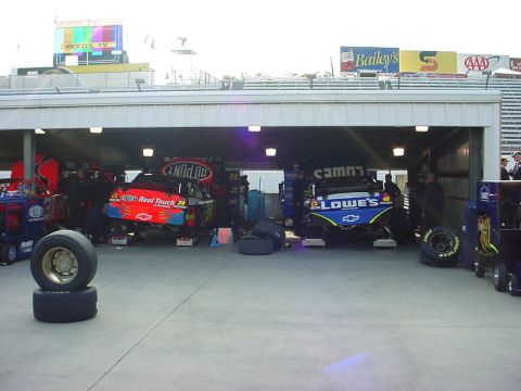 1993 Ford Mustang - Race cars are serviced in the pit area under a covered structure, with various sponsor logos visible on the vehicles and surrounding buildi…
