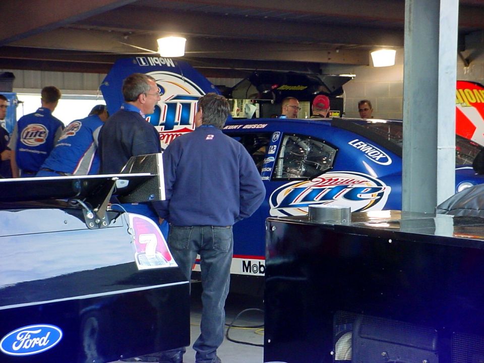 Two NASCAR race cars, one blue with Miller Lite sponsorship, the other dark with a Ford logo, in a garage setting