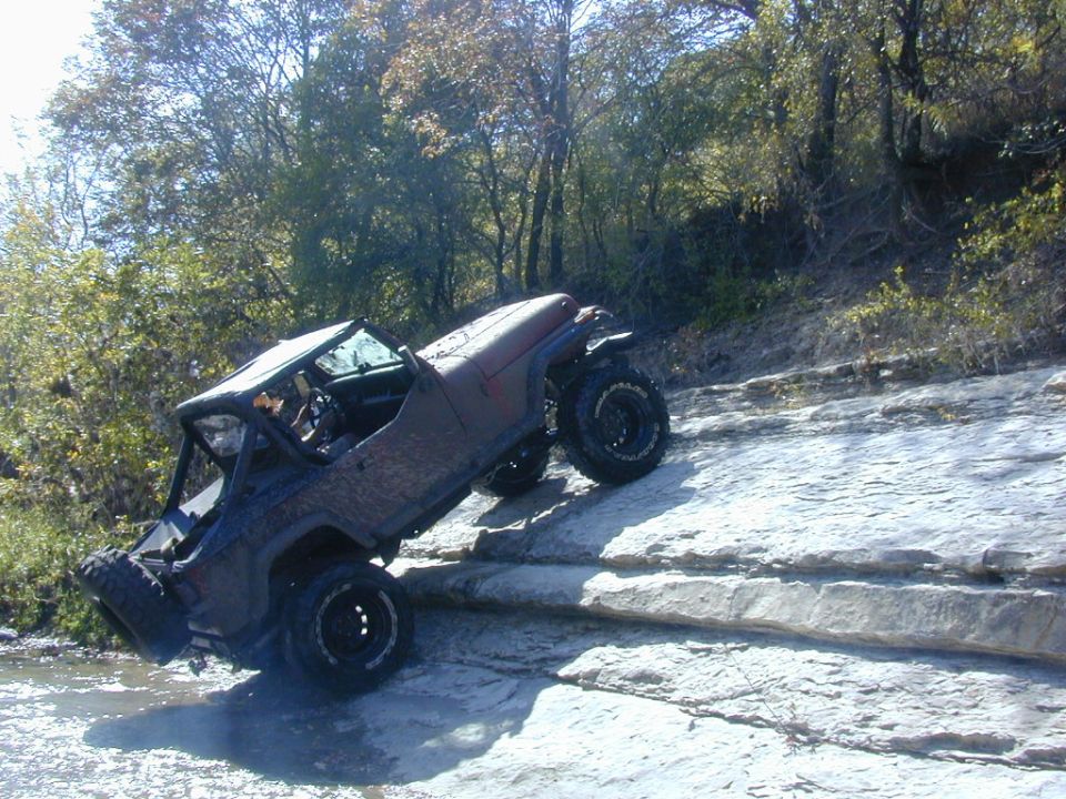 Lifted doorless red-primer Jeep crawling up a rock ledge on big off-road tires