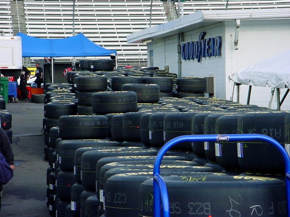 Stacks of Goodyear racing slicks in a track paddock beside a Goodyear building and grandstands