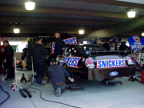 1993 Ford Mustang - Race cars with Snickers livery are being serviced in a garage pit area.