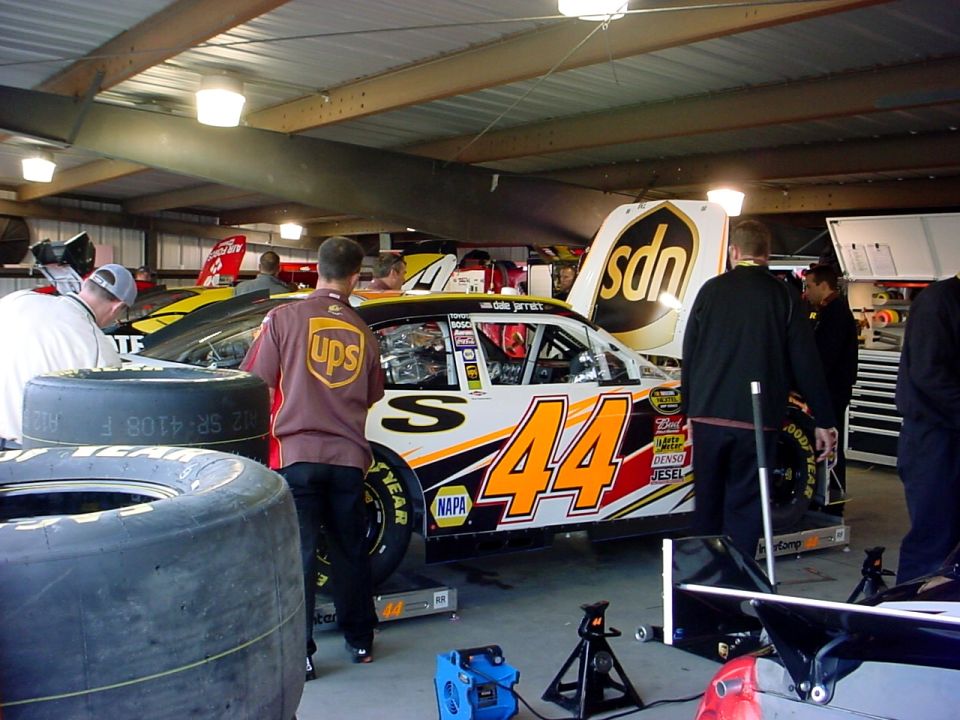 Race car number 44 with UPS livery in a garage, hood open, tires stacked nearby