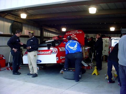 1993 Ford Mustang - A race car is being serviced in a garage by its pit crew, with the hood open and a tire being replaced.