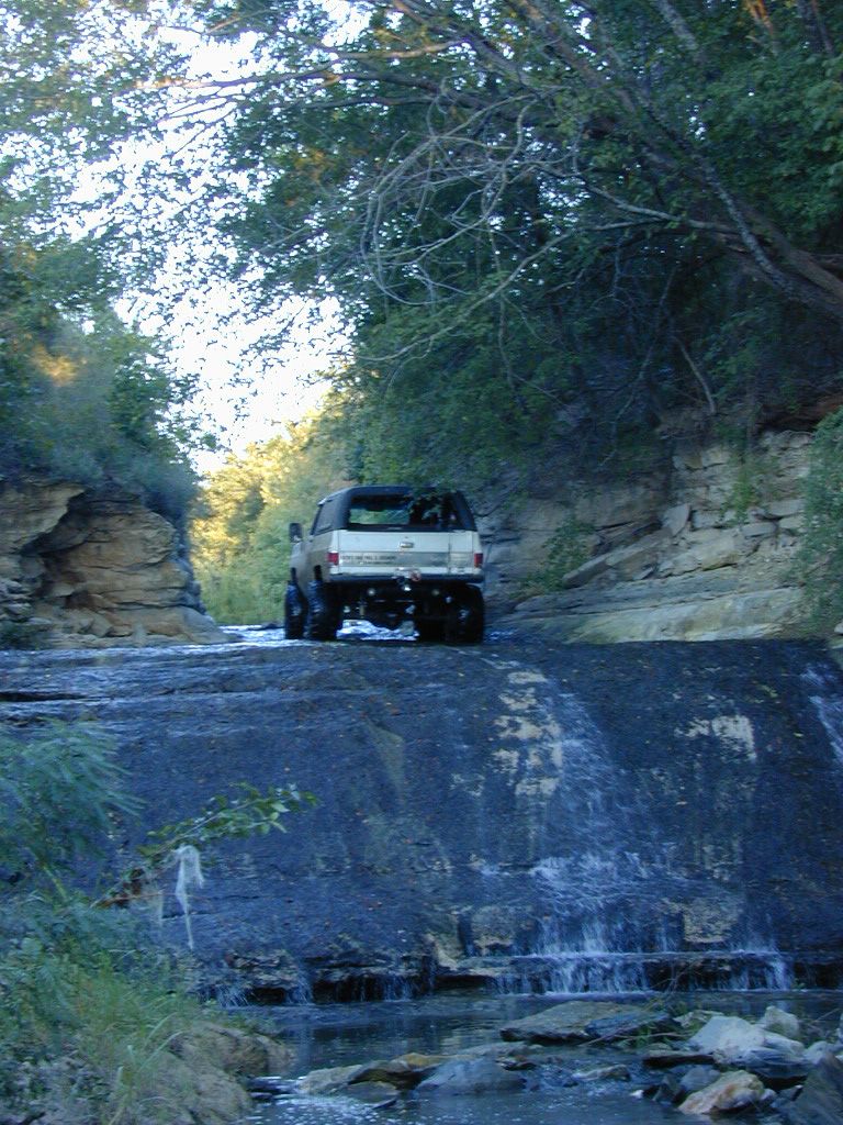 White lifted full-size SUV, rear view, on a rock ledge above a waterfall in the woods