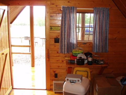 1978 Ford Bronco - The interior of a rustic cabin shows a table with camping gear and a view of the outdoors through a window and open door.