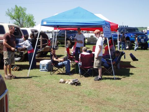 1978 Ford Bronco - Attendees relax under a canopy at an outdoor gathering, with various trucks parked nearby.
