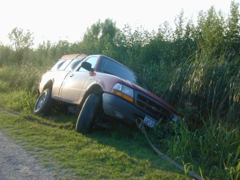 1996 Ford Bronco - A red pickup truck with a camper shell is stuck in tall grass, with a tow strap attached to the front.