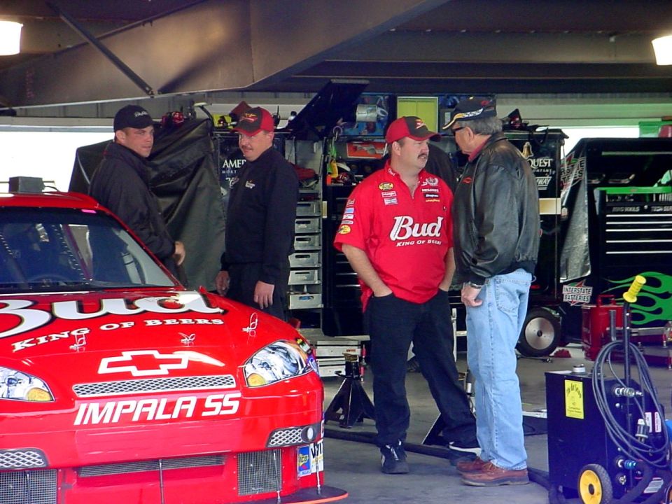 Red Budweiser Chevrolet Impala SS race car in a garage with crew members nearby