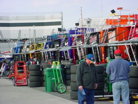 1993 Ford Mustang - A view of a race track pit lane, showing multiple garages and stacks of tires, with spectators in the background.