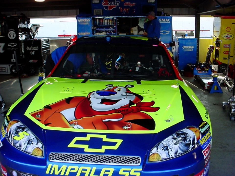 Race car with Tony the Tiger on the hood and 'IMPALA SS' on the front bumper, parked in a garage with pit equipment around