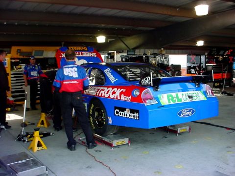 1993 Ford Mustang - A blue Ford race car is being serviced in a garage, with crew members around it.
