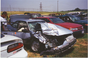 1996 Ford Bronco - A severely damaged silver sedan sits in a junkyard with its front end destroyed and driver's door ajar.