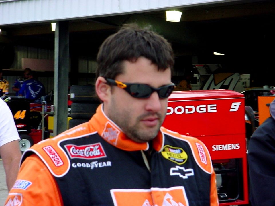 Driver in an orange-and-black firesuit with sunglasses near a Dodge race car