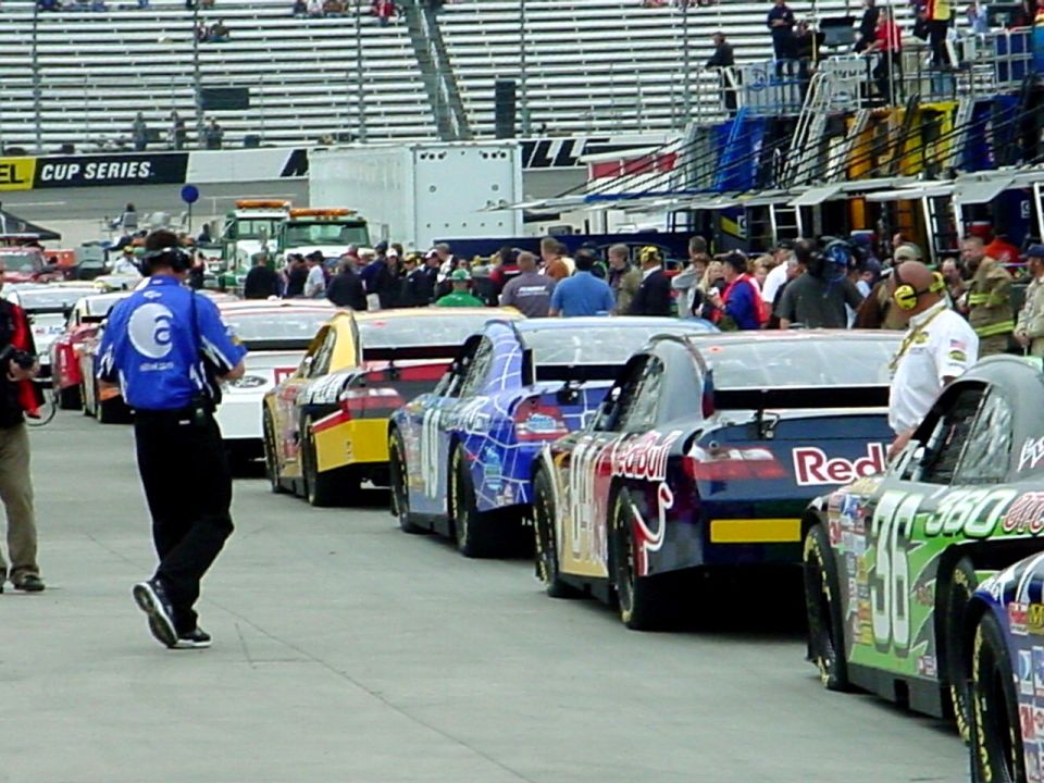 Several NASCAR race cars are lined up on a track, with pit crews and spectators in the background
