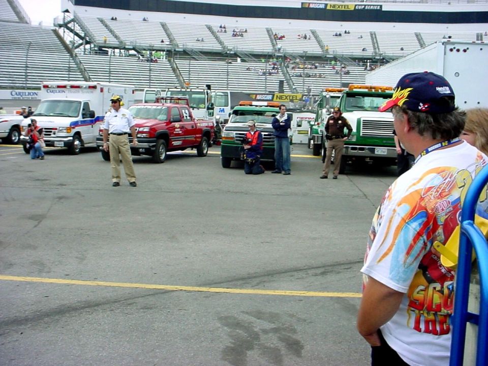 NASCAR pit area with tow trucks, an ambulance, and people standing around