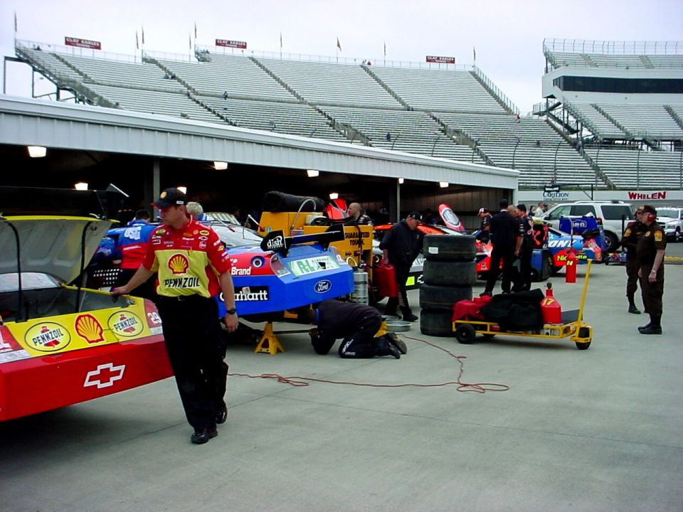 Race cars and crew in pit lane with grandstands in background