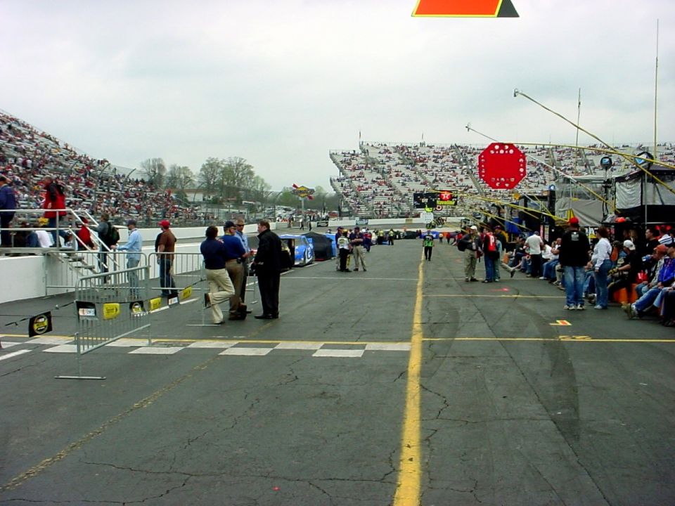 Race track with grandstands full of spectators, pit area with people, and a checkered finish line