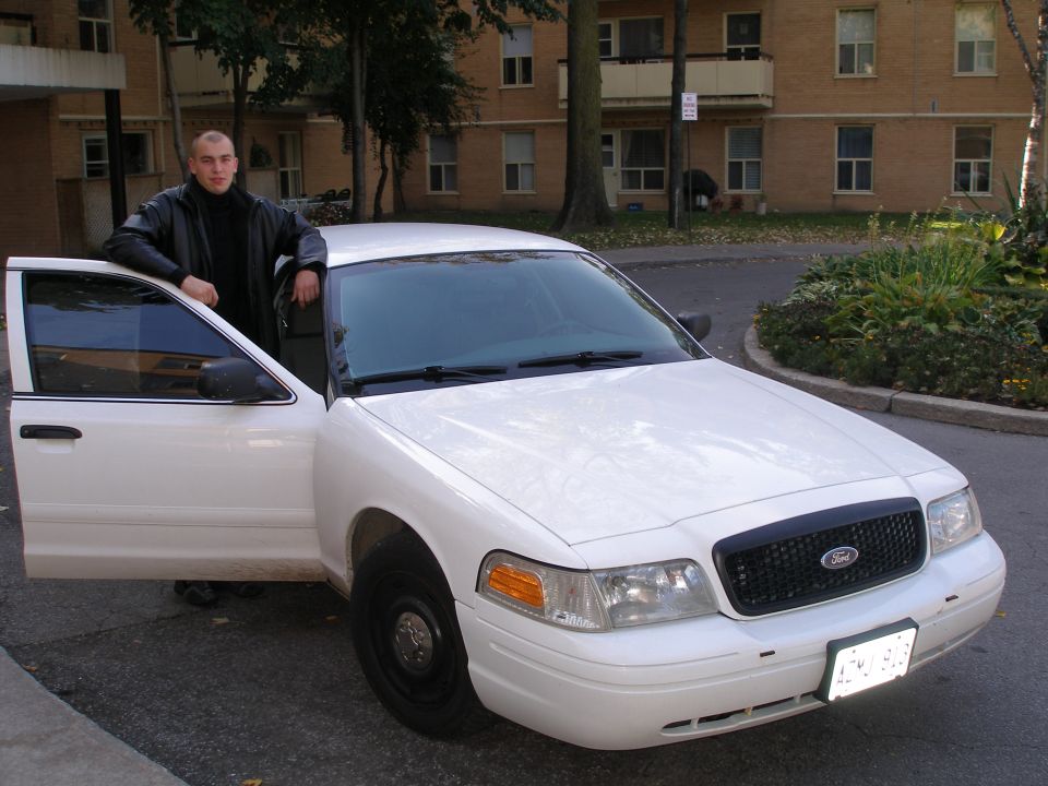 Man stands by a white Ford Crown Victoria with its driver's door open. The grille has a Ford logo