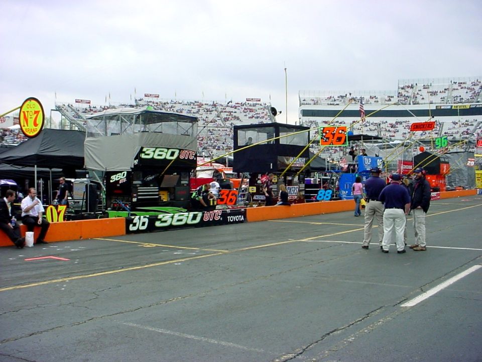 NASCAR pit lane with race cars, crew members, and grandstands filled with spectators