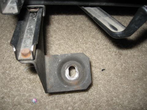 1990 Ford Bronco - A close-up view of a metal bracket with a hole, resting on a carpeted surface. A website logo is visible in the lower right corner.