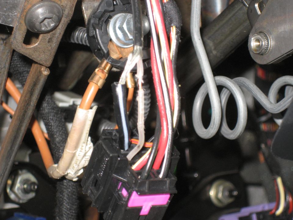 Under-dash wiring closeup with black connector, pink lock tab, crimp splice on orange wire, and taped harnesses