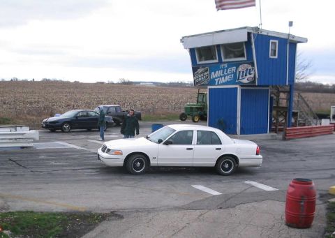 1998 Ford Crown Victoria - A white 1998 Ford Crown Victoria sedan is parked on a race track near a blue building with advertising.