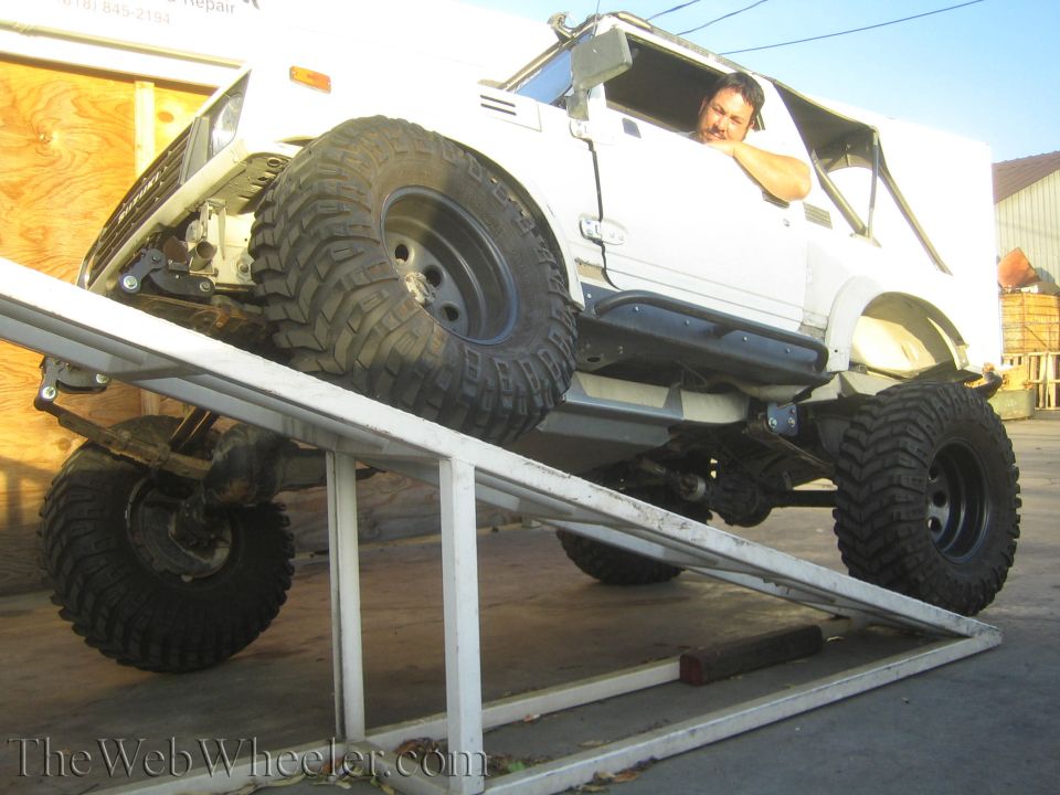1987 Suzuki Samurai with large off-road tires on a metal ramp, driver visible