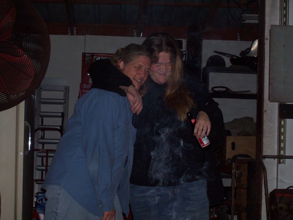 Two people embrace in a garage, one smoking. A large fan is to the left