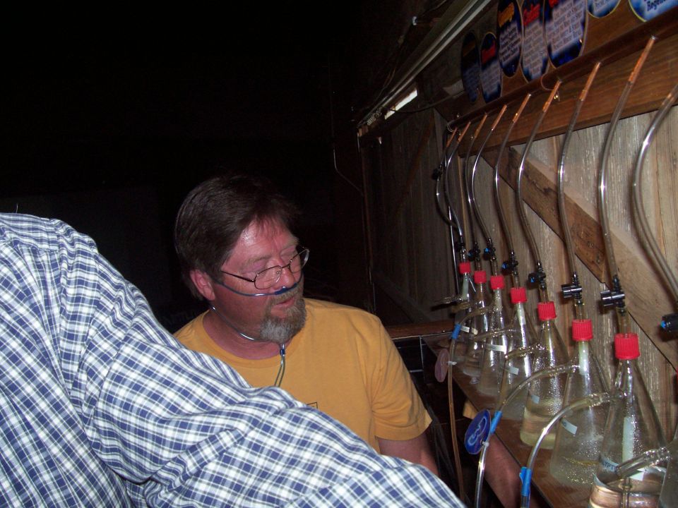 Man in glasses and yellow shirt next to a rack of glass carboys in a dim room