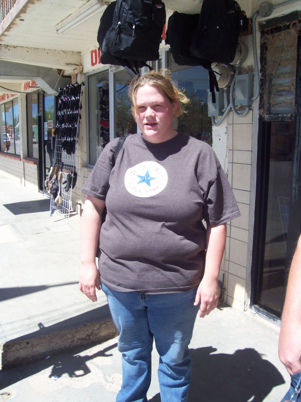 Woman in a gray t-shirt and blue jeans stands on a sidewalk in front of a store