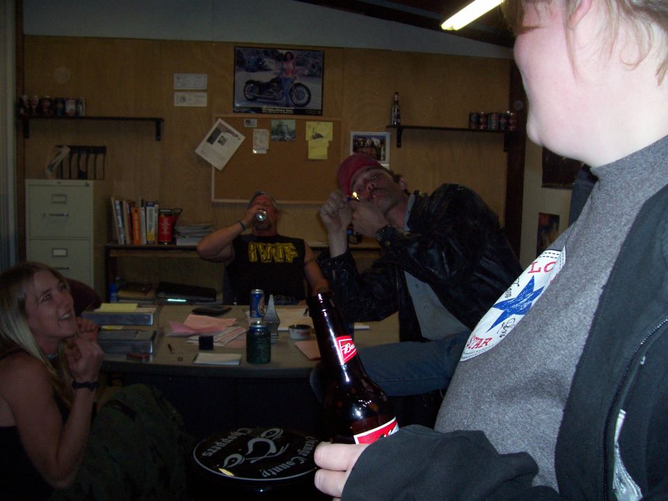 People gathered in an office, one lighting a cigarette, another drinking from a can