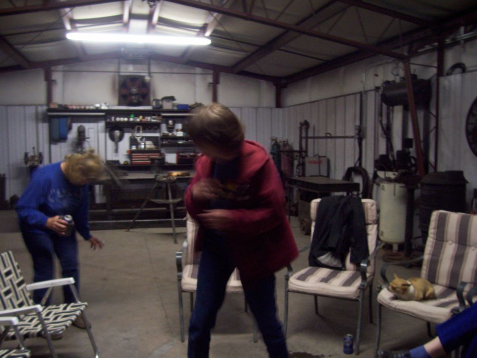 Garage interior with two people, tools, and chairs. A small dog rests on a chair