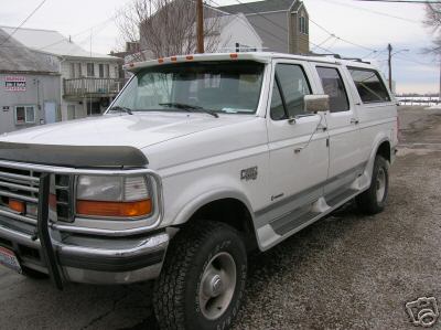 1995 Ford C-350 (Centurion Conversions) - This extended SUV features a brush guard and roof rack, parked on a gravel lot with buildings in the background.