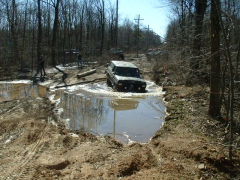 1991 Ford Bronco - An off-road vehicle navigates a challenging muddy terrain with trees and other vehicles nearby.
