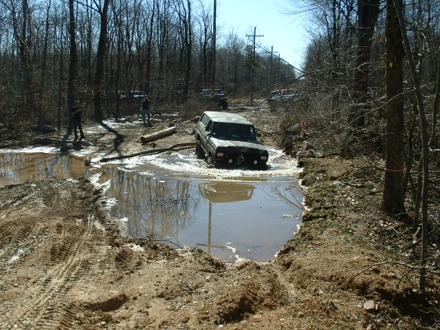 Ford Bronco driving through a muddy water obstacle in a wooded area. Other vehicles and people are visible in the background