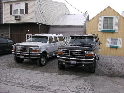 1995 Ford C-350 (Centurion Conversions) - Two Ford SUVs are parked in a gravel lot. The white SUV is a long wheelbase model, and the black SUV is in front.