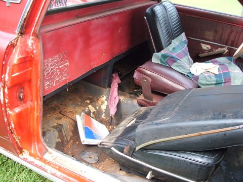1966 Ford Ranchero - The interior of a red vehicle shows significant rust on the floor and worn seats.