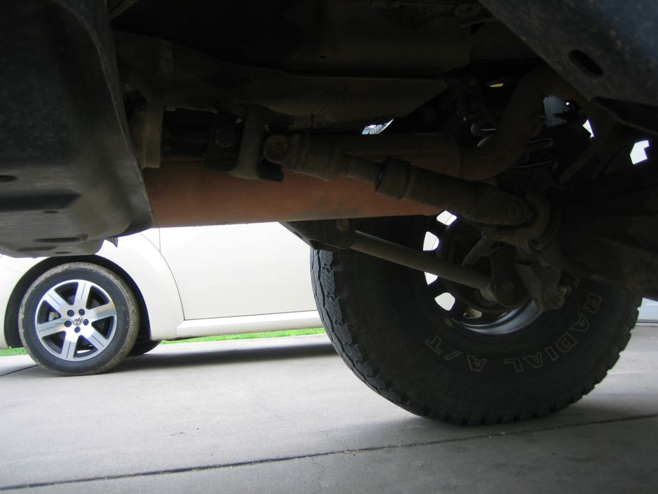 Underside view of a vehicle's drivetrain and tire, with another car visible in the background