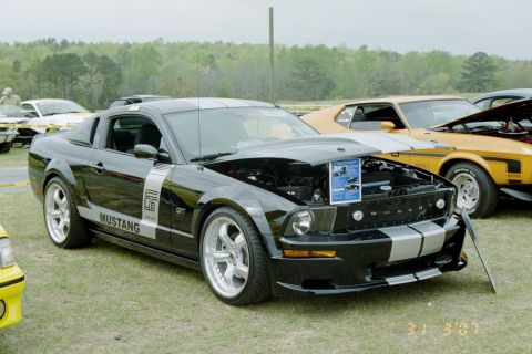 1976 Ford Ranchero - A black Ford Mustang Shelby GT is displayed with its hood open, parked on a grassy field with other vehicles nearby.