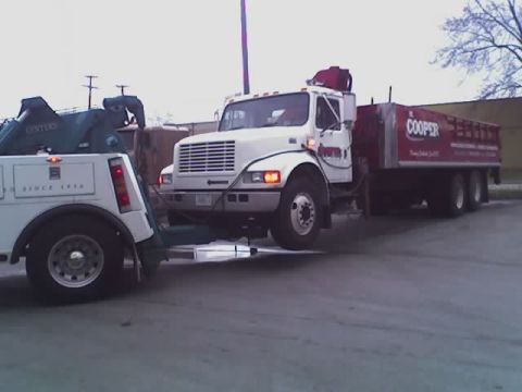 1992 Ford Bronco - A tow truck is seen towing a white flatbed truck that is pulling a red trailer with 'Cooper' branding.