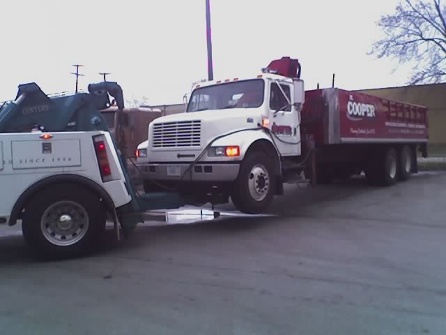 Tow truck towing a white flatbed truck with a red Cooper trailer