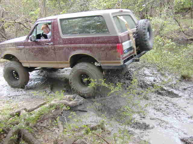 Maroon and tan Ford Bronco flexing out of a mud hole in the woods with a front wheel in the air