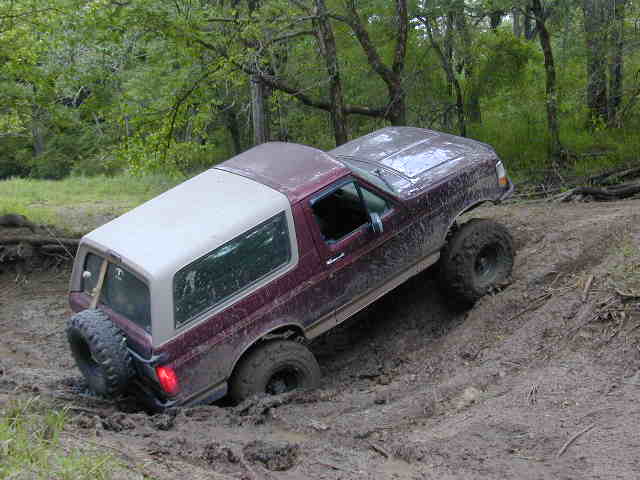 Burgundy Ford Bronco with white hardtop climbing out of a muddy rut in the woods