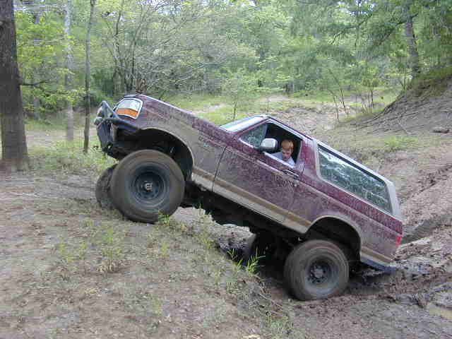 Muddy burgundy Ford Bronco pitched nose-up in a rut in the woods